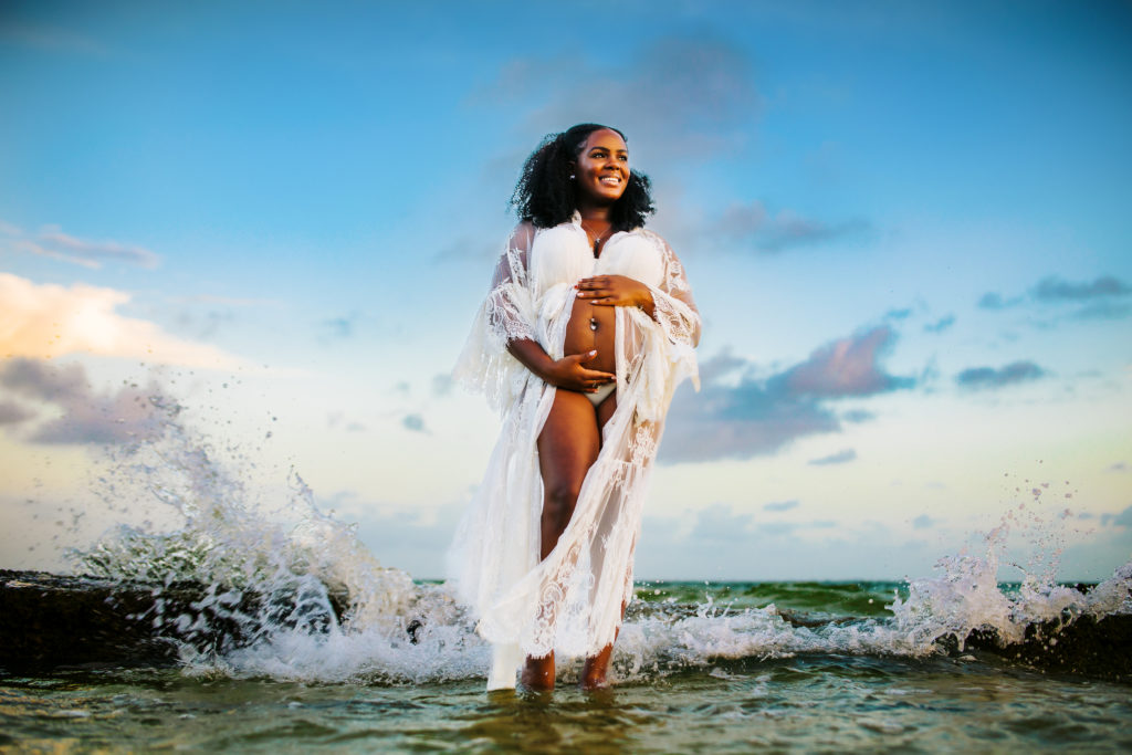 fun boho pregnancy announcement portraits taken at the beach in the water at sunset with a women in a lace robe holding her baby bump
