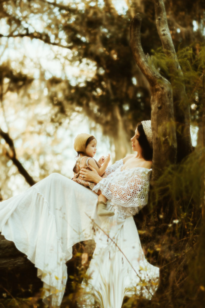 mother and daughter boho breastfeeding portraits with a women sitting on a fallen over tree talking to her 1 year old daughter in a dreamy woodsy park in tampa