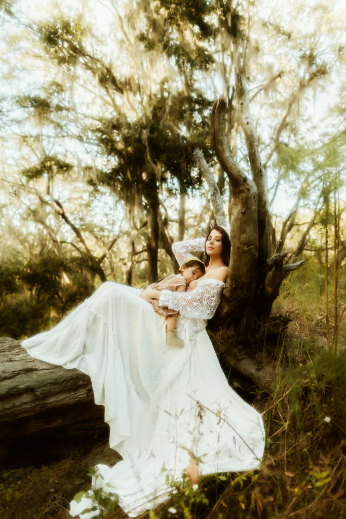 hippie retro boho breastfeeding portraits with a mom nursing her baby girl on a fallen over tree branch surrounded by greenery dressed in vintage clothing 