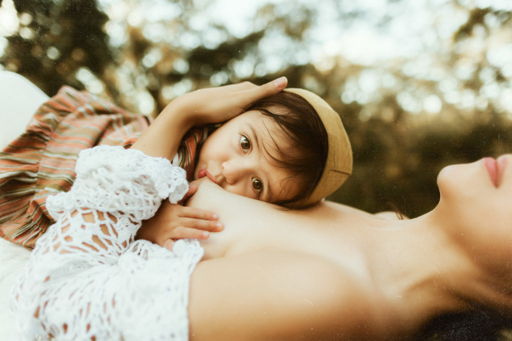 moody and dreamy boho breastfeeding portraits of a mom laying on a tree branch breastfeeding her 1 year old baby outdoors for a photo shoot 
