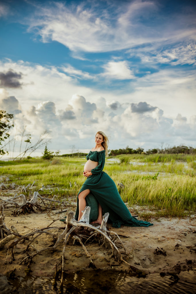whimsical boho maternity portrait of a pregnant women in a green boho skirt and top set standing in tall grass on the beach 