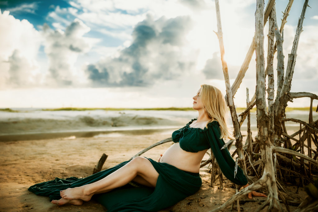 pregnant women sitting in the sand against a tree near a inlet of water at the beach cradling her baby bump during a maternity photo session