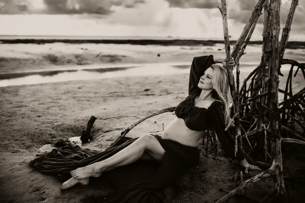 black and white beach pregnancy portrait of a women sitting in the sand looking up at the sky while birds fly around her