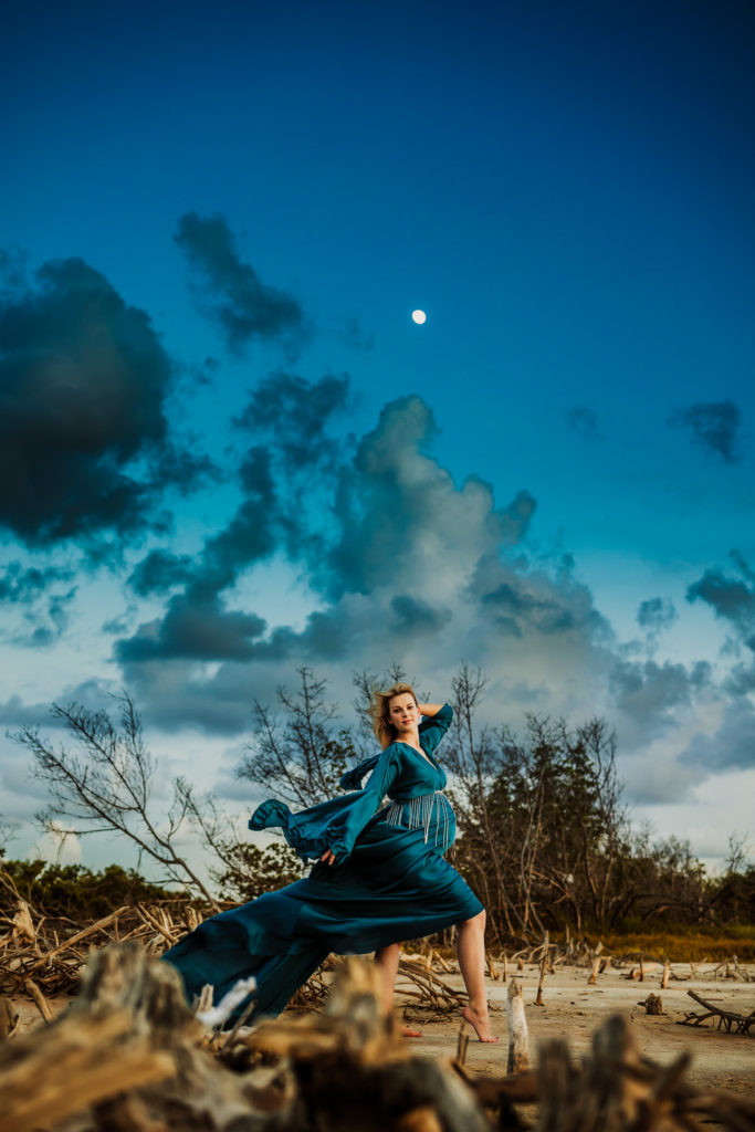 moody colorful maternity portrait of a women in a blue gown flying in the wind at the beach 