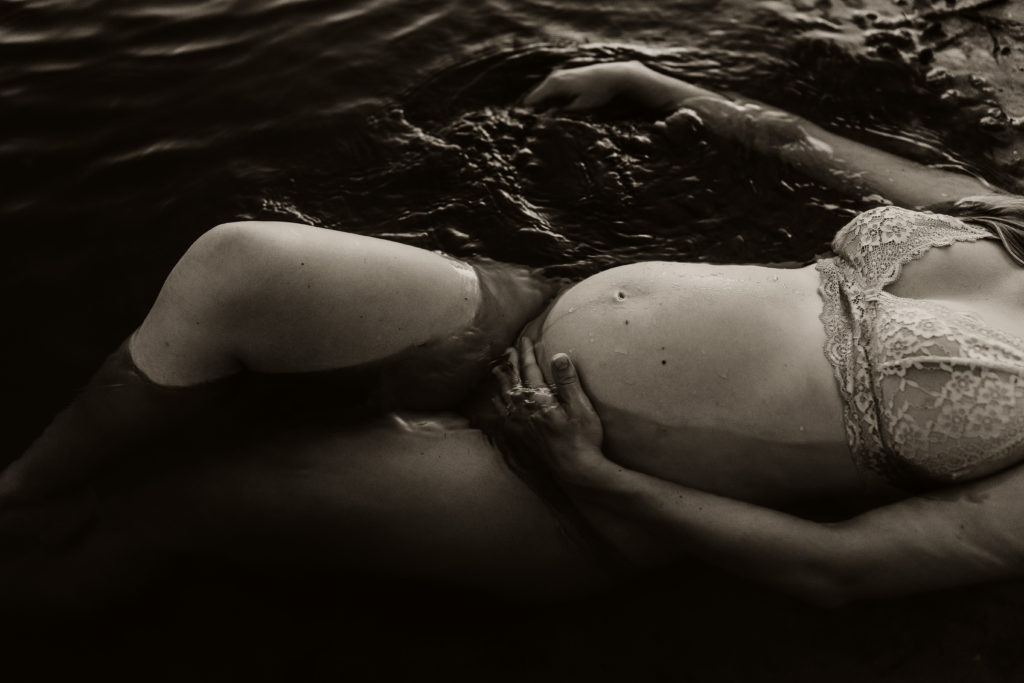 black and white pregnancy portrait at the beach shot from above with a women floating in the water holding her pregnant belly